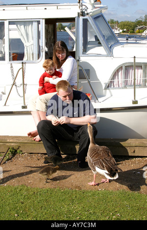 UK Norfolk Broads Horning Marina young holidaymaking family sat on steps of leisure craft feeding goose Stock Photo