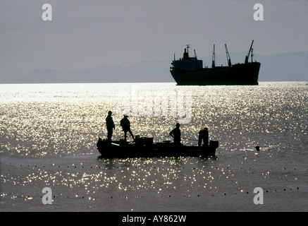 silhouettes of four men on small boat Aqaba harbour, Jordan. A large freight ship is anchored in the background Stock Photo