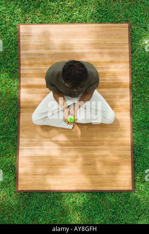 Man sitting crosslegged on mat outdoors, holding apple, high angle view Stock Photo