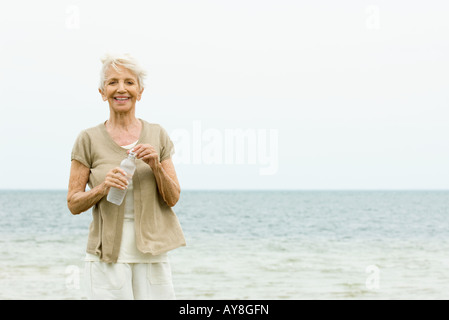 Senior woman standing at the beach, holding bottle of water, smiling at camera Stock Photo