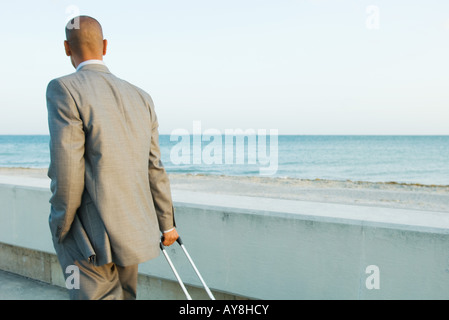Businessman walking by the sea, pulling suitcase, looking at view Stock Photo