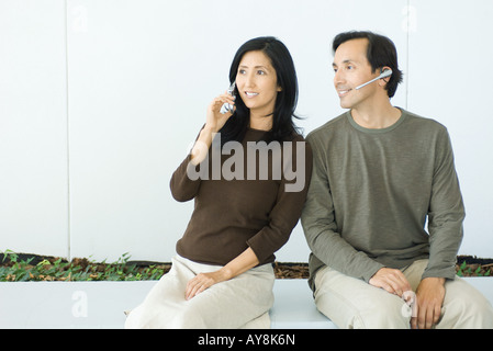 Couple sitting side by side, woman using cell phone, man wearing headset, both smiling Stock Photo