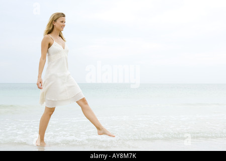 Young woman in sundress taking large step as she walks in shallow water on beach, full length Stock Photo