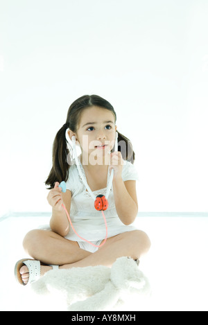 Little girl sitting next to teddy bear, listening to toy stethoscope Stock Photo