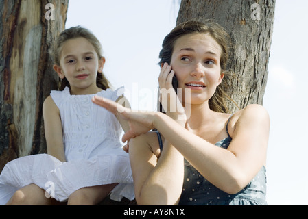 Young woman using cell phone, brushing little sister's hand off her shoulder