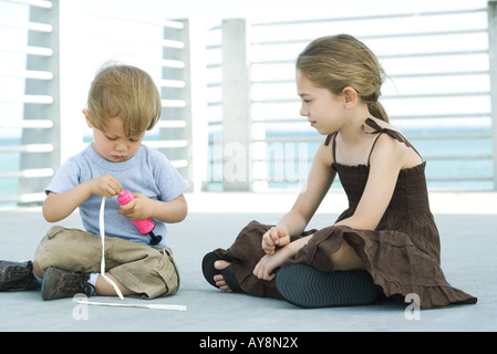 Little boy sitting on the ground inflating balloon with air pump, older sister watching