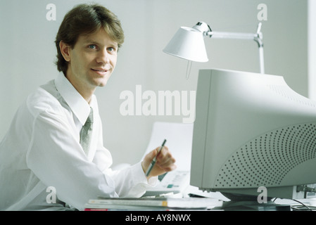 Businessman sitting at desk in front of computer, smiling, looking up Stock Photo