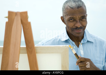 Senior man with hand under chin, holding paint brushes, smiling at camera Stock Photo