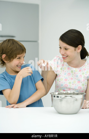 Brother and sister standing in kitchen, girl putting whipped cream on boy's nose, both smiling Stock Photo
