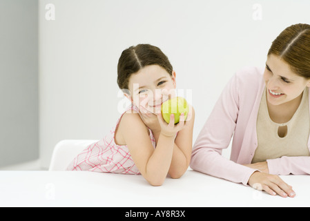 Little girl sitting at table with mother, hand under chin, holding apple, smiling at camera Stock Photo