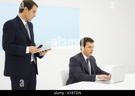 Two businessmen in office looking down at laptop computer together, one holding cell phone Stock Photo