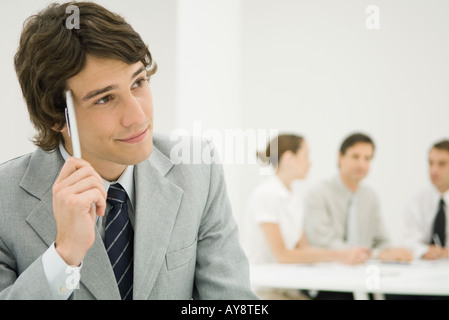 Businessman holding pen against temple, looking away, colleagues seated in background Stock Photo
