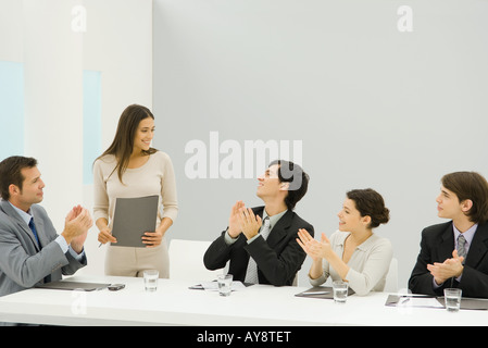 Businesswoman standing beside conference table, holding document, colleagues looking at her and clapping Stock Photo