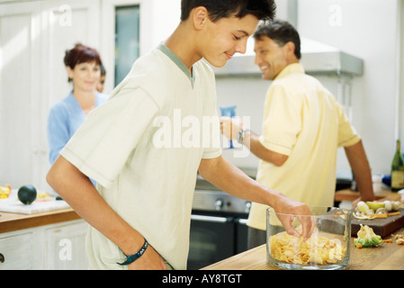 Teenage boy standing in kitchen, eating chips from bowl, family preparing food in the background Stock Photo