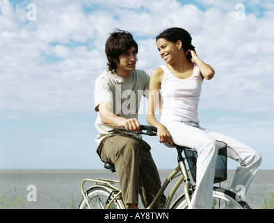 Young couple riding bicycle, female sitting on handlebars, looking over shoulder Stock Photo