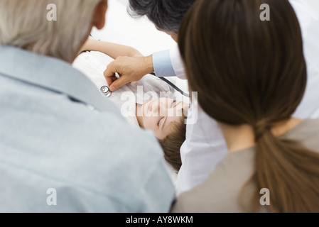 Doctor listening to boy's heart with stethoscope while family members watch, over the shoulder view Stock Photo