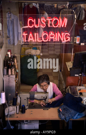 An elderly Hispanic woman works at sewing machine in the window of a custom tailor shop in New York City Stock Photo