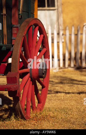 A red wagon wheel stands in front of a fading white picket fence, and an old adobe building, in Lincoln, New Mexico. Stock Photo