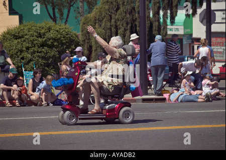 older woman riding electric wheeled chair scooter in parade waves at crowd on sidewalk Stock Photo
