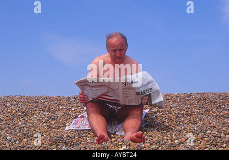 Middle aged man reading Times newspaper sport and business in swimming costume on beach Stock Photo