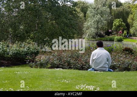 A man sitting on the grass relaxing in the rose gardens by a pond in Regents Park in London UK Stock Photo