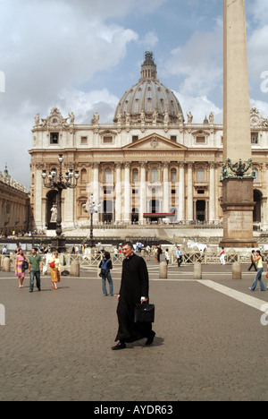 Priest wearing religious attire walking in St Peters Square backdrop of tourists and historical Basilica beyond on hot sunny day in Vatican Rome Italy Stock Photo