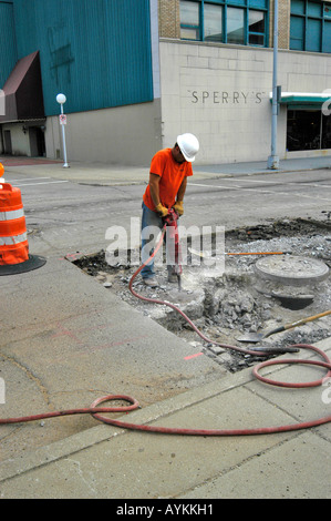 A construction worker wearing a safety helmet stands using a jackhammer to remove old cement that is under asphelt Stock Photo