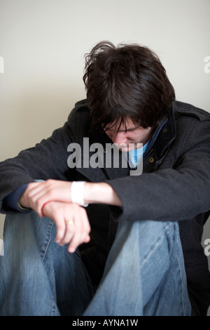 young dark haired teenage man sitting on the floor with back against the wall in the fetal position looking down Stock Photo