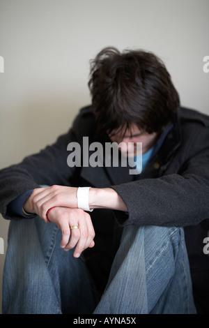 young dark haired teenage man sitting on the floor with back against the wall in the fetal position looking down Stock Photo