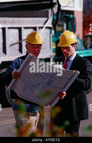 Caucasian architect and construction foreman on the job site wearing hardhats and looking at blueprints Stock Photo