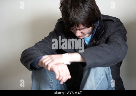 young dark haired teenage man sitting on the floor with back against the wall in the fetal position looking down Stock Photo