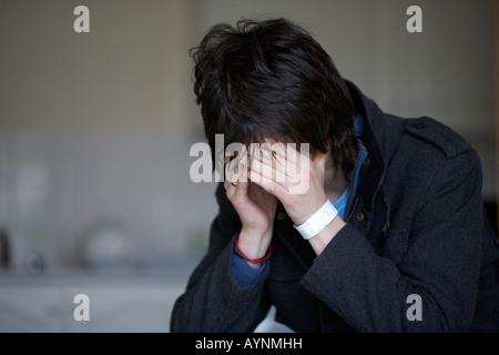 young dark haired teenage man sitting with his head in his hands hiding his face staring at the floor in the kitchen Stock Photo