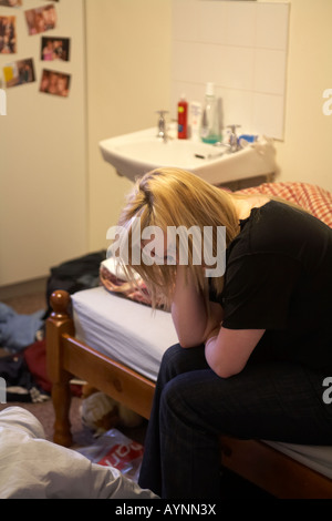 young teenage woman sitting on bed in messy bedsit bedroom holding her head in one hand Stock Photo