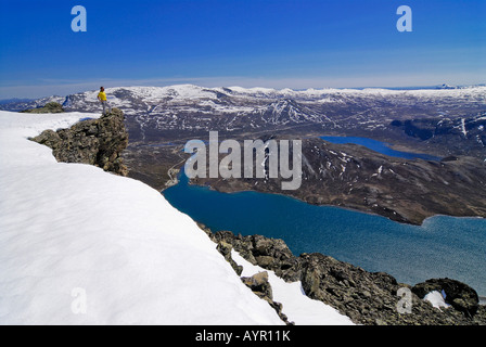 Hiker standing on a snow-covered rock pulpit looking over a vast landscape with mountains and lakes, Jotunheimen National Park, Stock Photo
