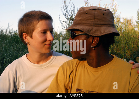 Mixed couple sitting outdoors during golden hour, woman with short hair looking at man wearing a brown bucket hat and sunglasses. Stock Photo