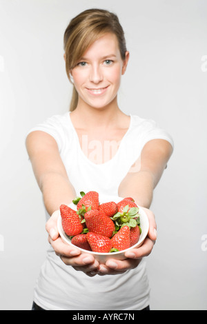 Smiling young woman with long dark-blonde hair offering a bowl of fresh strawberries Stock Photo