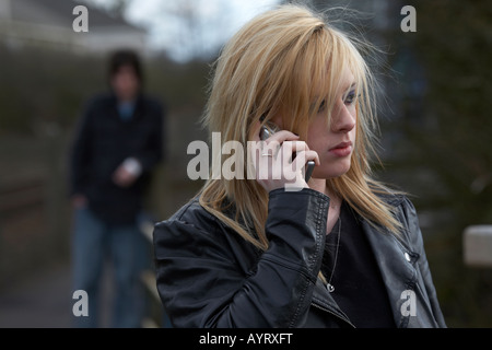 blonde haired teenage woman on mobile phone making a call standing with man standing watching close behind woman being stalked social distancing Stock Photo