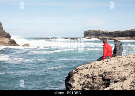 Father and son sat on rocky ledge in bay looking out to sea Stock Photo