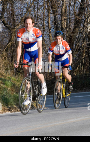 Two college aged young male cyclists climbing a hill on a bicycle in team kit / jersey on road. Stock Photo