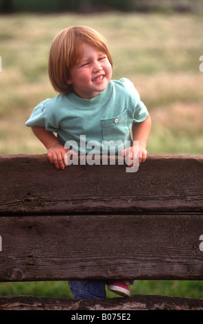 A smiling child with red hair climbs a wooden fence. Stock Photo