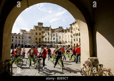 a group of cyclists in a tour party leave Piazza Anfiteatro Romano in Lucca Tuscany Stock Photo