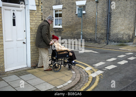 Elderly gentleman (husband) with wife in wheelchair negotiating the small step of a path on street Stock Photo