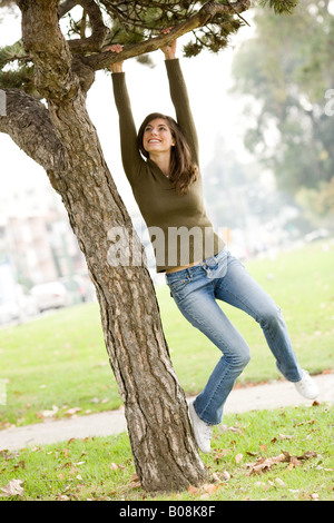 A woman jumping for joy, laughing, smiling in a park and playfully swinging from a tree limb. Stock Photo