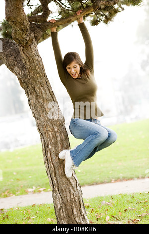 A woman jumping for joy, laughing, smiling in a park and playfully swinging from a tree limb. Stock Photo