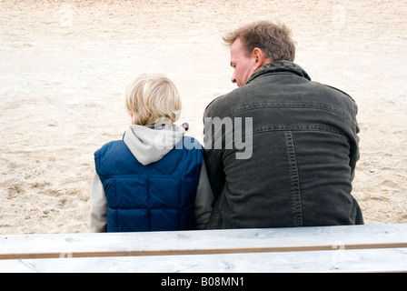 A father looks lovingly at his blonde haired son while they are sitting on a bench during a day out at the beach. Stock Photo