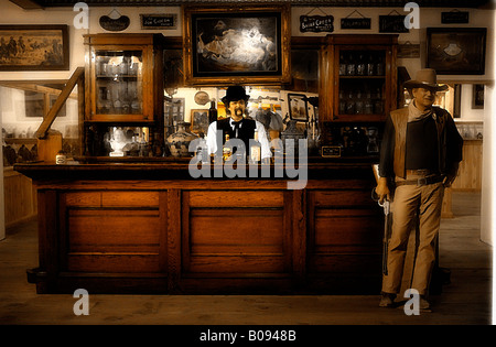 Image of a manikin bartender behind a vintage western saloon counter with a cardboard figurine of John Wayne standing near. Stock Photo