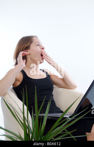 young woman with laptop sitting in an armchair, yawning Stock Photo