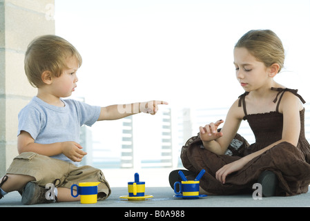 Siblings sitting on the ground together, brother pointing a sister, sister looking down at finger Stock Photo