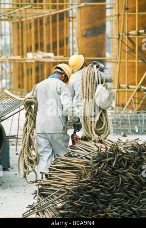 Workers at construction site carrying rope, metal rods stacked in foreground Stock Photo