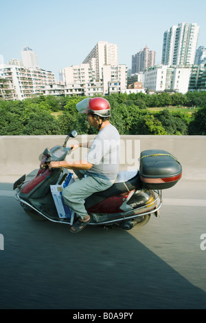 China, Guangzhou, man riding motor scooter, high-rise buildings in background, side view Stock Photo